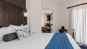 Quarto de hotel com cama grande, cabeceira de madeira, mesa de cabeceira com abajur, vista para uma lareira e janela com cortinas leves.