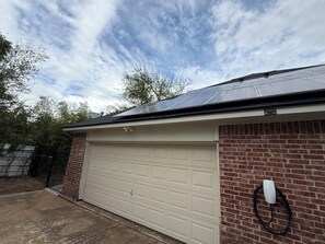 Un garage à l’extérieur en briques, avec une porte de garage blanche et un toit équipé de panneaux solaires.