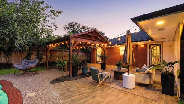Um pátio nos fundos com pérgola de madeira, mobiliário de jardim e luzes pendentes.