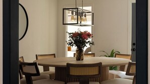 Uma sala de jantar com uma mesa redonda, seis cadeiras, um lustre e um vaso com flores.