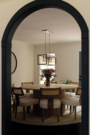 A dining area with a round table, six chairs, a hanging light fixture, and a vase with flowers. - Designer Pool Retreat (Houston)