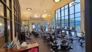 Un gimnasio con diverso equipamiento de ejercicio, amplias ventanas y una pantalla de televisión.