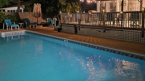 Piscina con sillones de descanso, una terraza de madera y un área cubierta con luces colgantes.