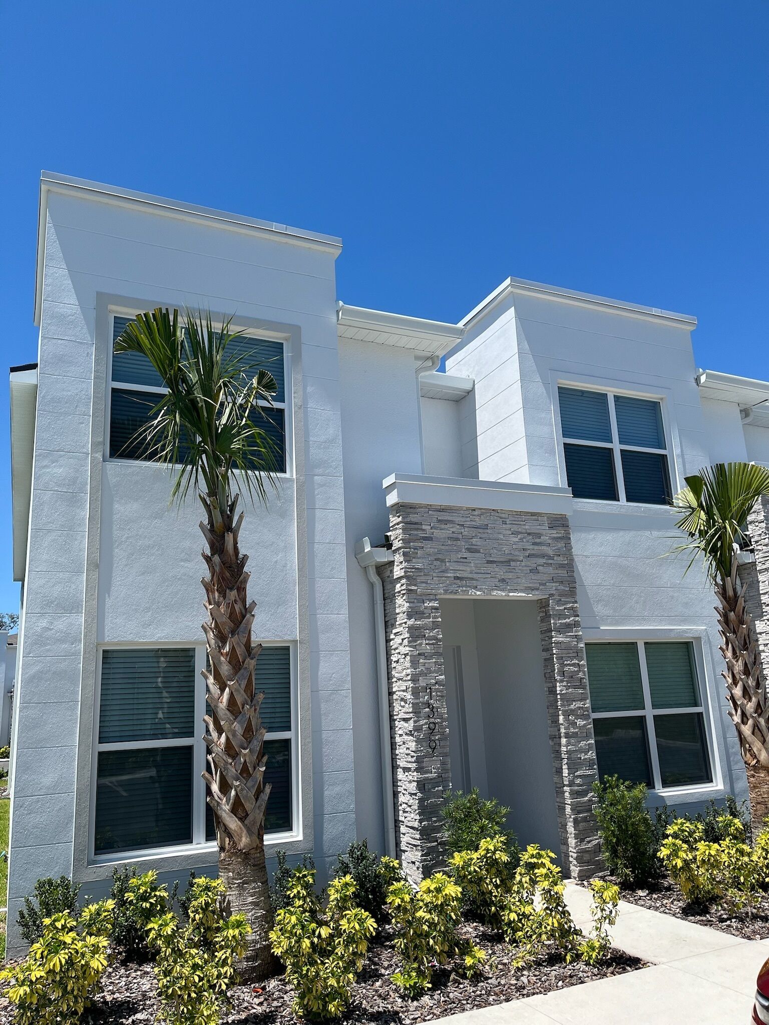 A modern two-story house with a stone facade, large windows, and a well-maintained garden with palm trees and shrubs.