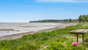 Ein Strand mit einer Holzbank, Rasenflächen und einer entfernten Küste mit Gebäuden.