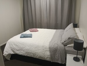 Un lit bien fait, recouvert d’une couette blanche, d’une couverture grise et de deux serviettes pliées, une bleue et une rose.