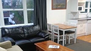 Un salon avec un canapé en cuir noir, une table basse en bois, un fauteuil beige, une table à manger avec des chaises blanches, un coin cuisine avec un four à micro-ondes et un tableau accroché au mur.