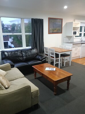 A living room with a black leather sofa, a wooden coffee table, a beige armchair, a dining table with white chairs, a kitchen area with a microwave, and a painting on the wall.