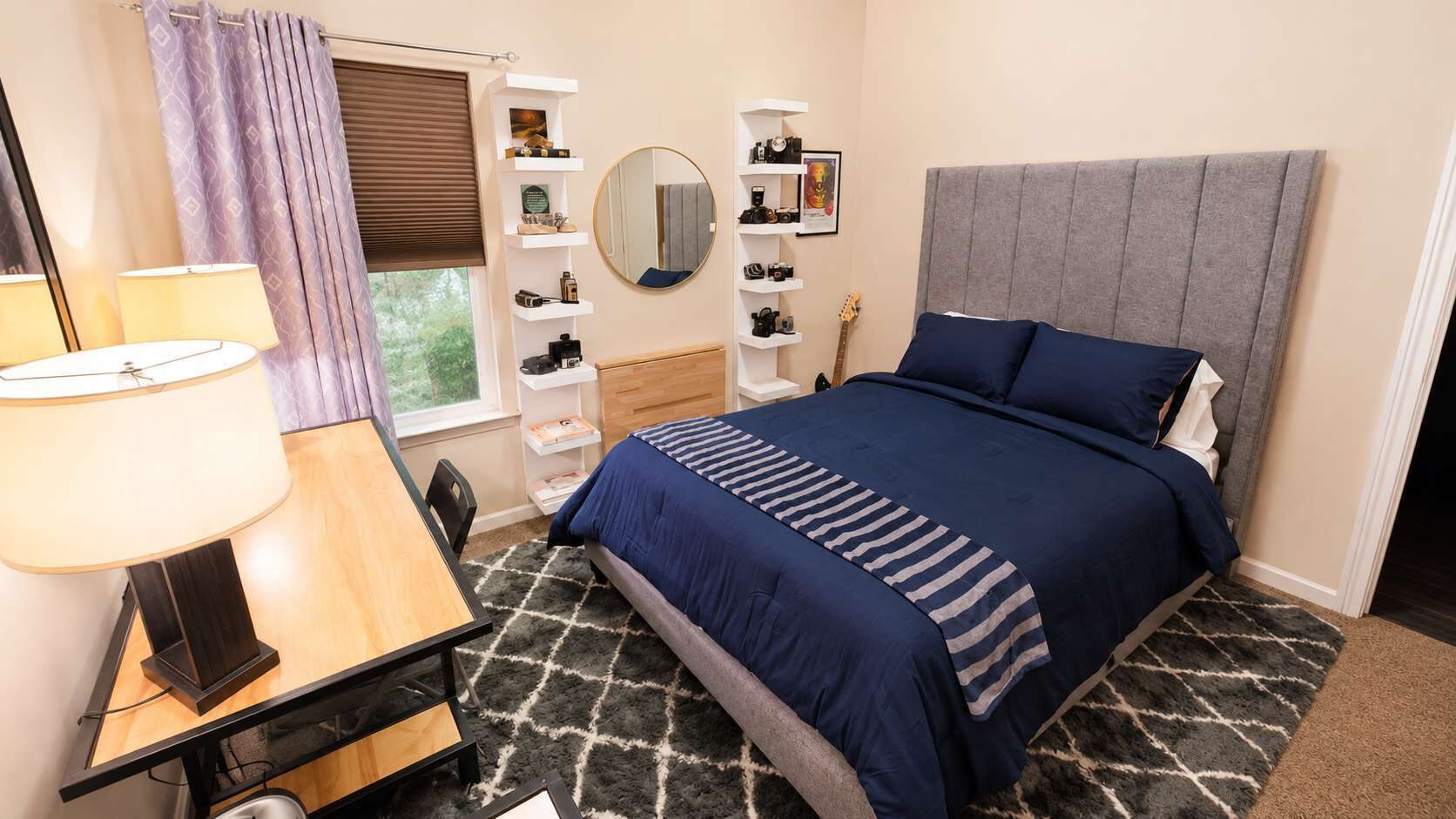 Une chambre à coucher comprenant un lit, des tables de chevet, un bureau, une lampe, un miroir et une fenêtre avec des rideaux.