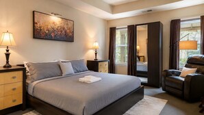 A modern bedroom with a large bed, bedside lamps, a reclining chair, and a window with curtains.