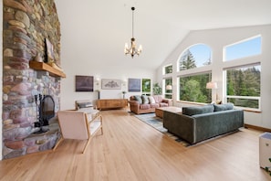 Ein geräumiges Wohnzimmer mit Kamin aus Stein, einer Kristallleuchtre, einem Sofa, einem Couchtisch und großen Fenstern mit Blick ins Freie.