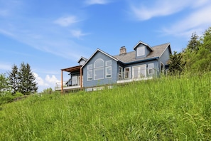 A two-story house with a large front porch, a covered area, and a grassy hill surrounding it. - Washougal 4BR - Hot Tub, Sauna, Private 5 Acres (Washougal)
