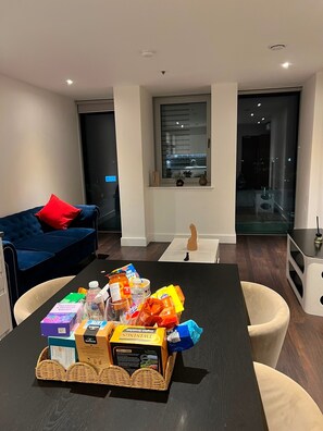Een moderne woonkamer met een donkerblauwe bank, een klein tafeltje met lekkernijen, een rieten mand en uitzicht op de stad door een groot raam.