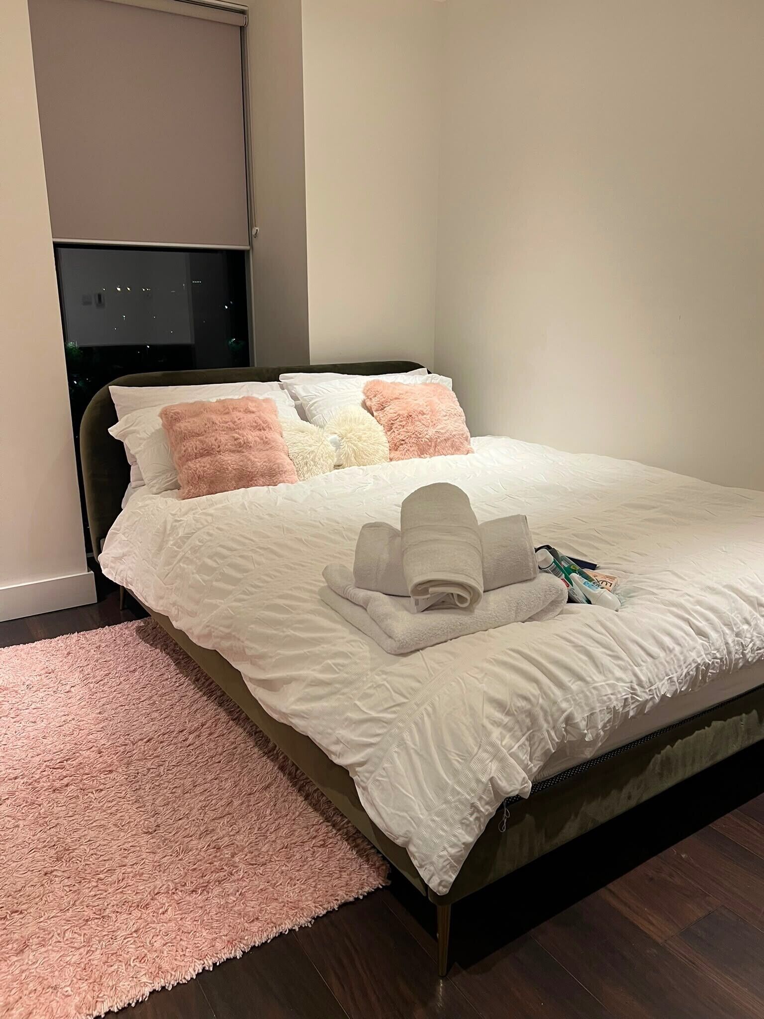 Een net opgemaakt bed met witte beddengoed, een roze pluizig vloerkleed en een zwart hoofdbord.