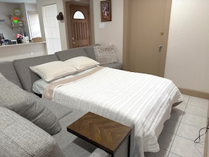 Quarto com cama, sofá cinzento, mesa de cabeceira de madeira e uma porta.