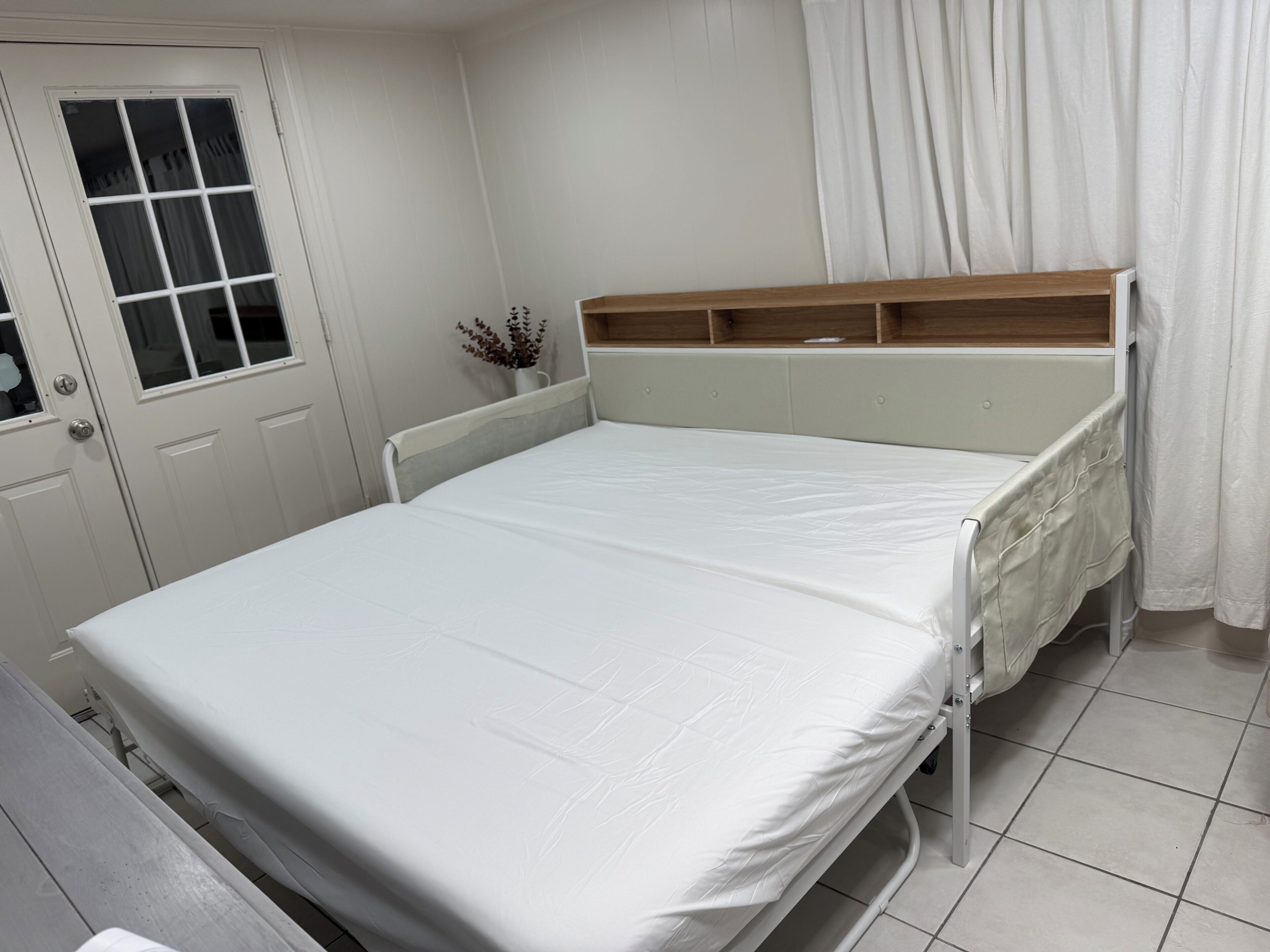 A single-bed room with a wooden headboard, a bedside table, a window with curtains, and a door with a window.