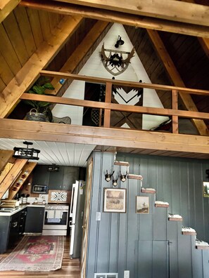 A loft-style room with a sloped ceiling, wooden beams, a kitchen area, and a dining table.