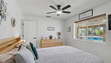 Een slaapkamer met een bed, een plafondventilator, een dressoir en een raam met uitzicht op een zwembad.
