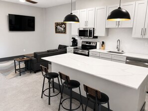 Een moderne keuken met witte kasten, een groot eiland, zwarte barkrukken en een flatscreen televisie aan de muur.