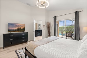 Ein Schlafzimmer mit einem Bett, einem Fernseher auf einer Kommode, Blick auf einen Balkon mit Gartenmöbeln und einer Tür zu einem weiteren Raum.