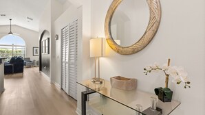 Een modern interieur met een glazen console tafel, een grote spiegel, een lamp en een vaas met witte bloemen.