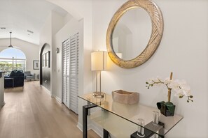 Ein modernes Interieur mit einem Glastische, einem großen Spiegel, einer Lampe und einer Vase mit weißen Blumen.