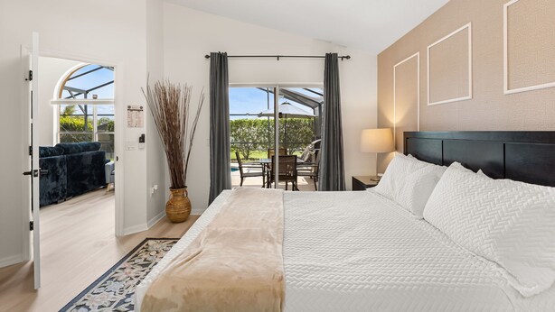Een slaapkamer met een groot bed, een nachtkastje, een lamp, uitzicht op een balkon en een deur naar een andere kamer.