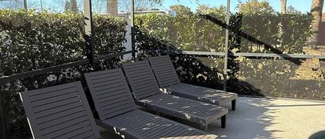 A covered outdoor seating area with wooden lounge chairs and a concrete floor.