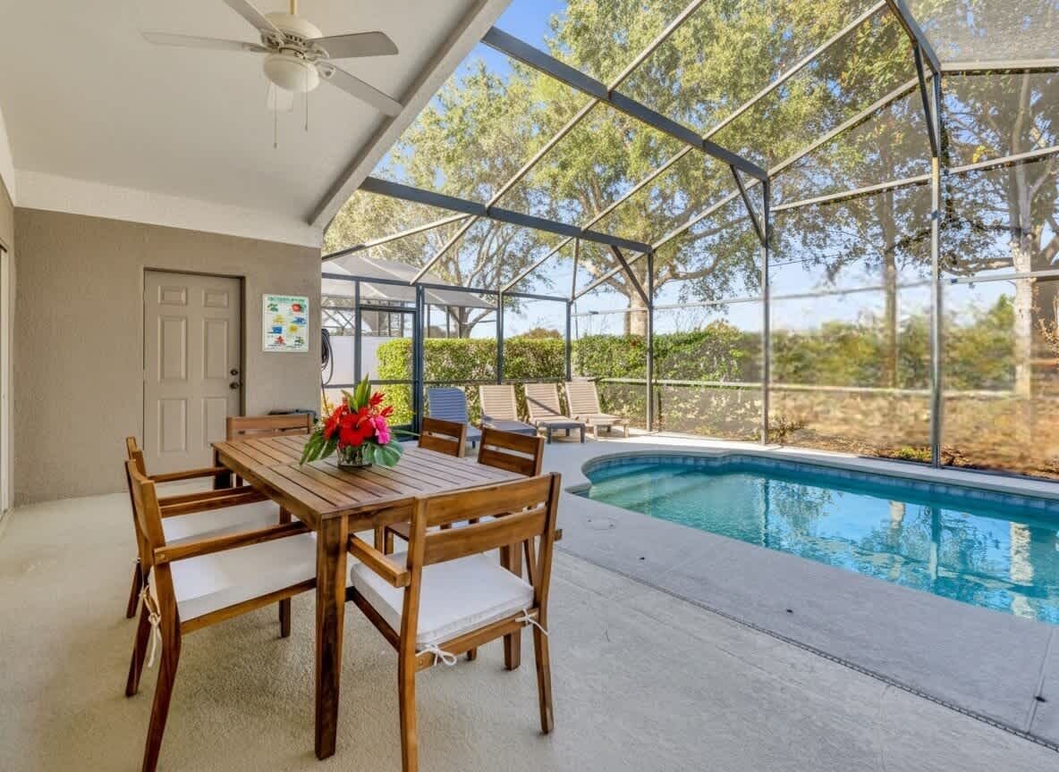 A covered outdoor area with a pool, wooden table and chairs, and a ceiling fan.