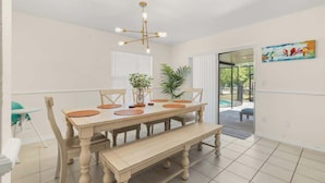 Un espace repas comprenant une table en bois, des chaises, un banc, un lustre, des plantes en pot et une porte-fenêtre coulissante donnant sur un espace extérieur.