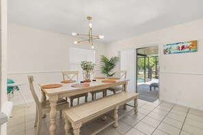 A dining area with a wooden table, chairs, a bench, a chandelier, potted plants, and a sliding glass door leading to an outdoor area.