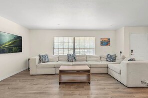 A living room with a sectional sofa, a coffee table, a large window with blinds, and artwork on the walls.