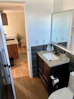 A modern bathroom with a granite countertop sink, dark wood cabinets, a large mirror, and a toilet paper holder. - Upscale, Family Friendly Coastal Retreat.
2 Blocks from So Cal’s, Seal Beach (Seal Beach)