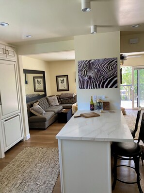 A modern kitchen with a marble island, a dining area with a black chair, and a living room with a grey sofa.