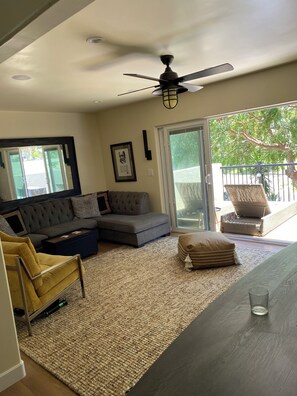 A living room with a sectional sofa, a yellow armchair, a glass coffee table, a ceiling fan, and a view through a sliding glass door to an outdoor area.