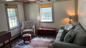 Una habitación con un sillón estampado, un sofá, una mesita y una lámpara. Hay dos ventanas con persianas, una alfombra estampada y vigas de madera a la vista en el techo.