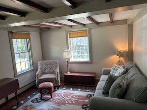 A room with a patterned armchair, a sofa, a small table, and a lamp. There are two windows with blinds, a patterned rug, and exposed wooden beams on the ceiling.