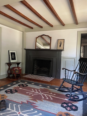 A room with a fireplace, a rocking chair, a patterned rug, and wooden beams on the ceiling.