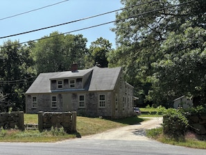 A two-story stone house with a gabled roof, a stone wall entrance, and a gravel driveway.