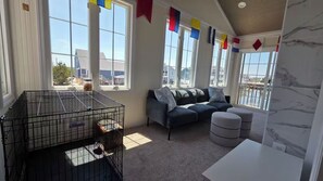 A room with a large window, a sofa, ottomans, a table, and a birdcage. - Mallard Cove | 170' Waterfront, Pool & Hot Tub (Stafford Township)