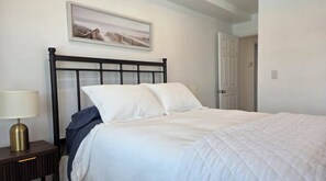 Una cama bien hecha en una habitación de diseño sencillo, una mesita de noche con una lámpara y una puerta a la derecha.