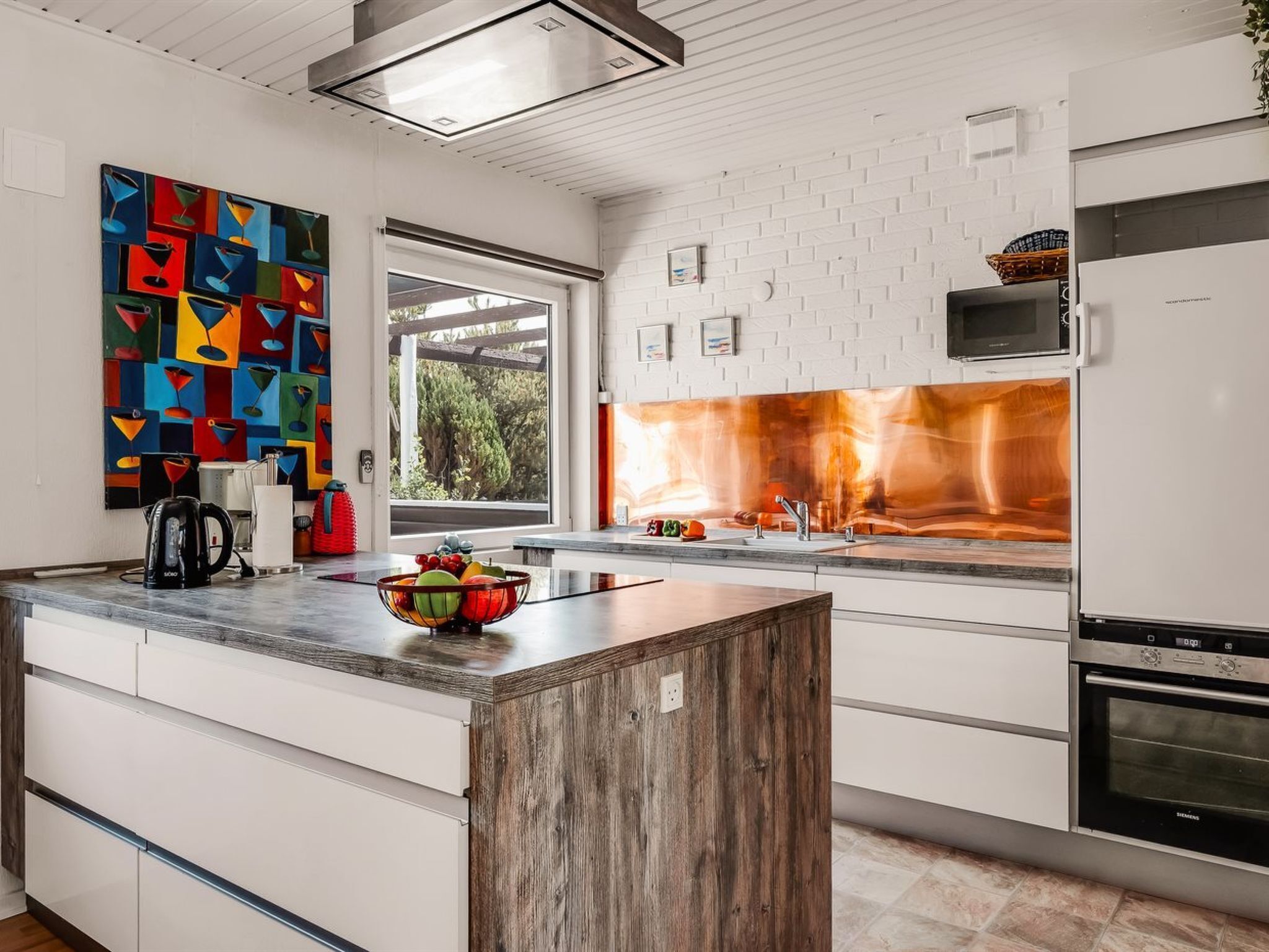 Une cuisine moderne avec des meubles blancs, un îlot central en bois, une peinture abstraite aux couleurs vives et une vue par la fenêtre.