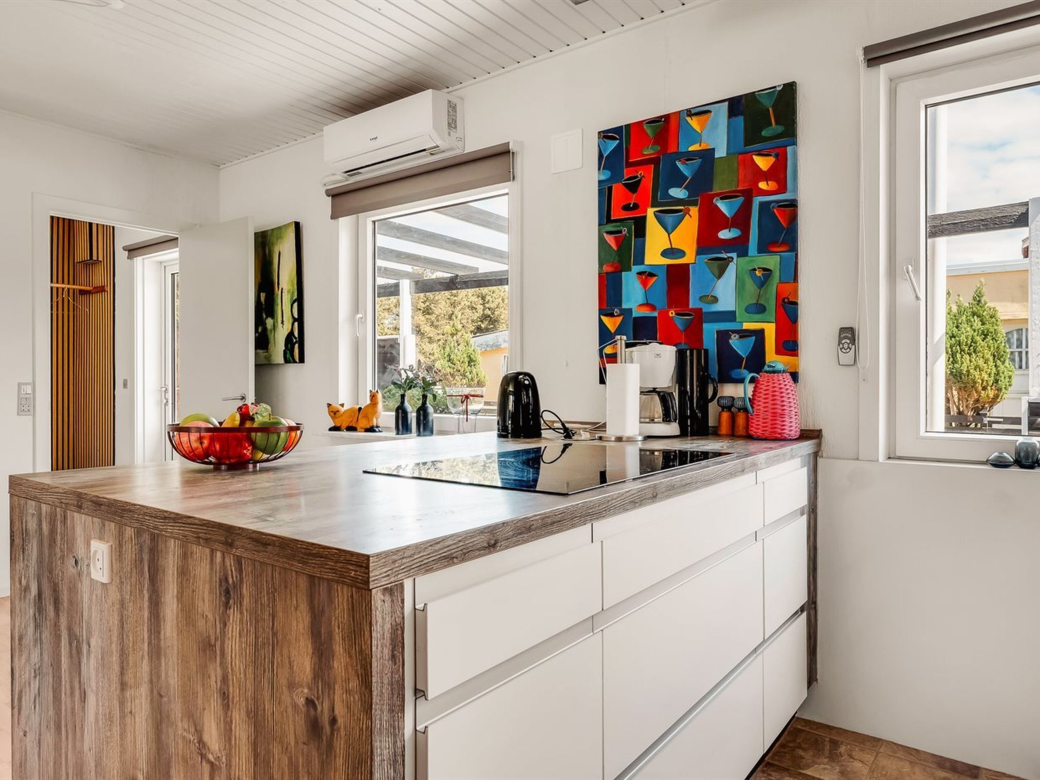 A modern kitchen with a wooden island, white cabinetry, a colorful abstract painting, and a view through a window.