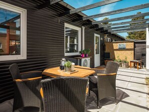 Een overdekt terras buiten met rieten meubels, een houten tafel en potplanten.