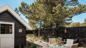 Eine Terrasse mit Korbstühlen, einem Glastisch, einem kleinen Gartenhaus und einem Holzzaun.