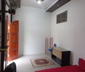 Una habitación con puerta de madera, un escritorio pequeño, una silla, cama roja, una obra de arte en la pared y una alfombra estampada.