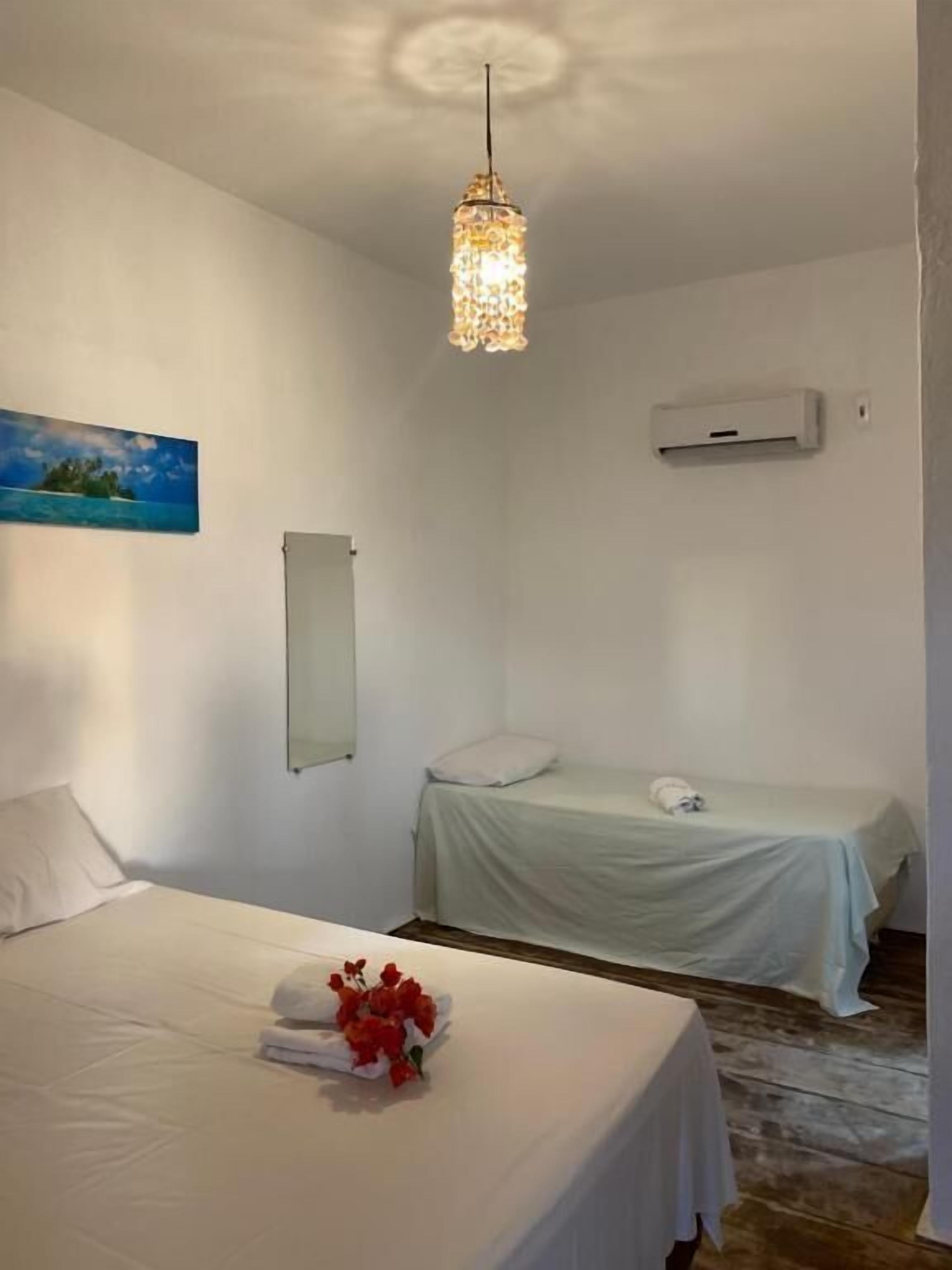 A hotel room with a bed, a bedside table with a flower arrangement, a hanging light fixture, a painting on the wall, and an air conditioning unit.