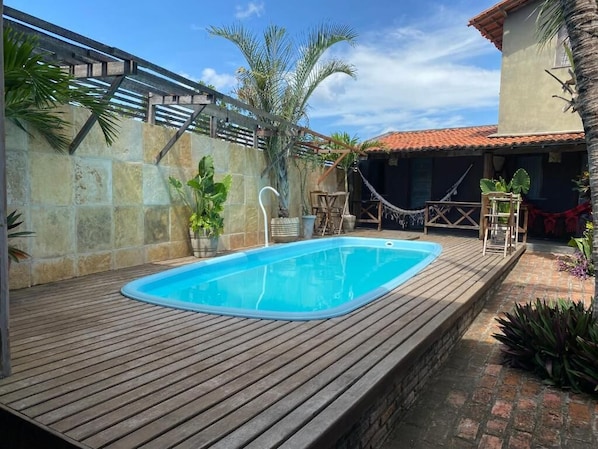 A rectangular swimming pool with a wooden deck, surrounded by stone walls and palm trees. - Pousada Kalango (Cajueiro da Praia)