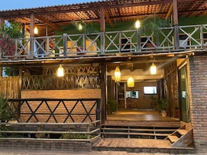A two-story building with a wooden balcony, outdoor seating area, and hanging lights. - Pousada Kalango (Cajueiro da Praia)