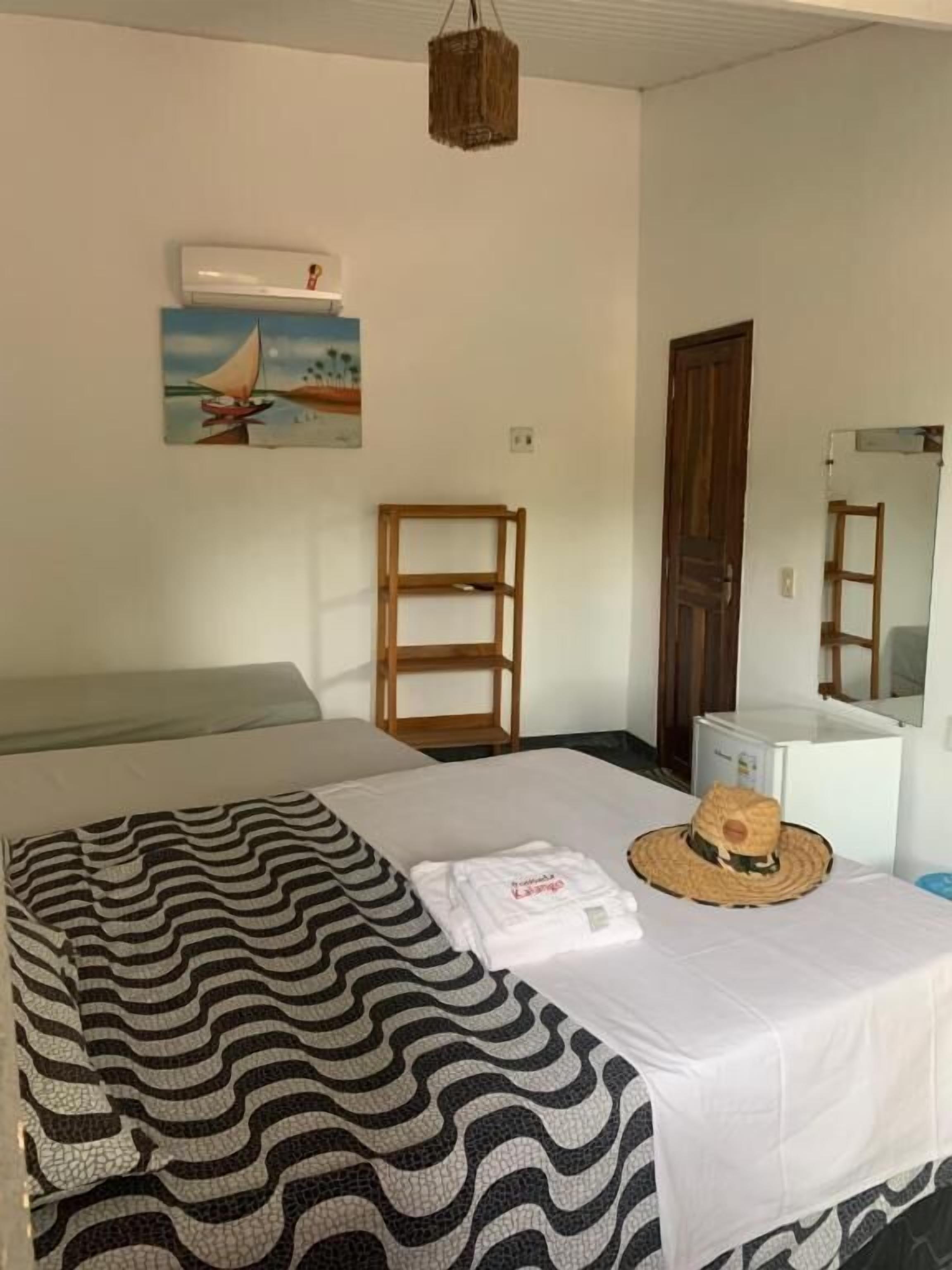 Un dormitorio con cama, mesita de noche con un sombrero, una silla, estante de madera, aire acondicionado blanco y un cuadro en la pared.